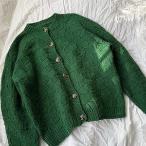 May include: A green cardigan sweater with a textured, woven pattern. The sweater has a round neckline, long sleeves, and a button-up front with five brown buttons. The garment is made of a thick knit material.