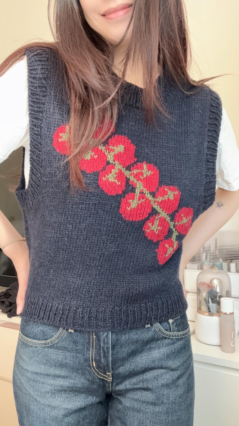 May include: A navy blue knitted vest featuring a vibrant red tomato design with green stems. The sleeveless top is worn over a white t-shirt and paired with blue denim jeans. The vest's design adds a pop of color to the outfit.