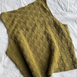 Op de afbeelding: Een mouwloze gebreide top in een gedempte olijfgroene kleur met een getextureerd dambordpatroon, gecreëerd door afwisselende vierkanten van tricotsteek en ribbelsteek over het gehele oppervlak van het kledingstuk met een eenvoudige ronde hals.