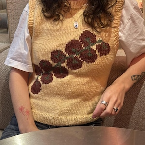 May include: A yellow knitted vest with a floral design in shades of burgundy and green. The vest is worn over a white t-shirt. The person is wearing rings and a necklace.