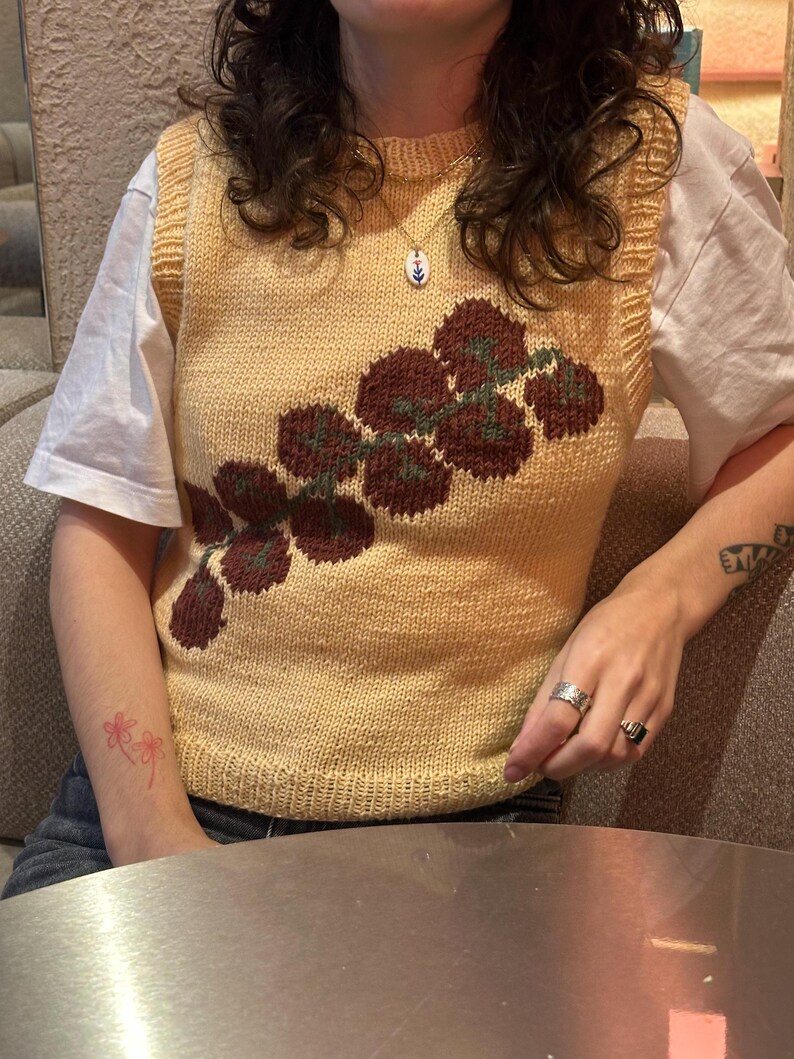 May include: A yellow knitted vest with a floral design in shades of burgundy and green. The vest is worn over a white t-shirt. The person is wearing rings and a necklace.