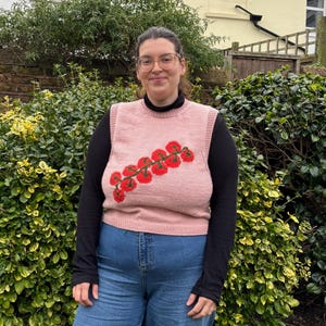 May include: A light pink knitted vest with a red and green tomato design. The vest is worn over a black long-sleeved shirt and paired with blue jeans. The person is standing in front of a green hedge.