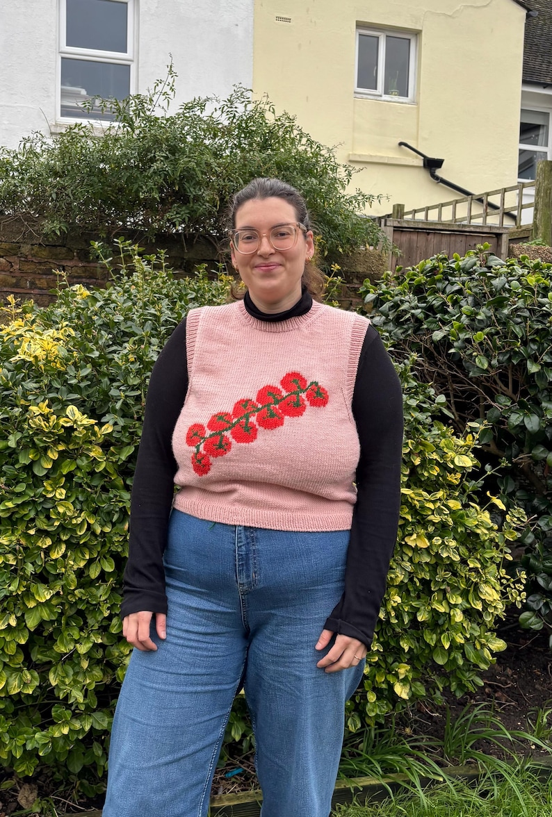 May include: A light pink knitted vest with a red and green tomato design. The vest is worn over a black long-sleeved shirt and paired with blue jeans. The person is standing in front of a green hedge.