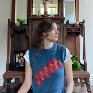 May include: A blue knitted vest with a red and green tomato design. The sleeveless top is worn over a white t-shirt. The vest is a crew neck style. The background includes a wooden cabinet and a green rabbit figurine.