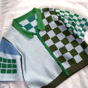 May include: A handmade crochet cardigan in a patchwork design. The cardigan features a checkerboard pattern in green and light blue, with solid light blue and blue sections. It has a green collar, button closure, and trim.