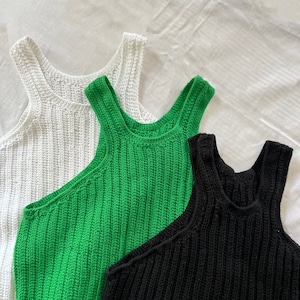 Puede incluir: Tres camisetas sin mangas de punto acanalado en blanco, verde y negro. Las camisetas sin mangas tienen cuello redondo y un diseño ajustado. La tela parece ser un punto de peso medio. Las camisetas se muestran sobre una superficie blanca.