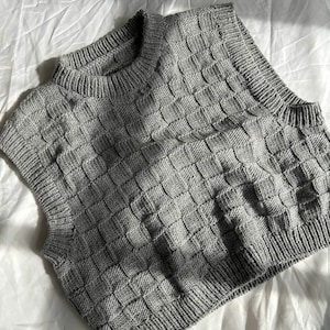 Könnte beinhalten: Ein grauer, ärmelloser Strickpullover mit quadratischem Muster.