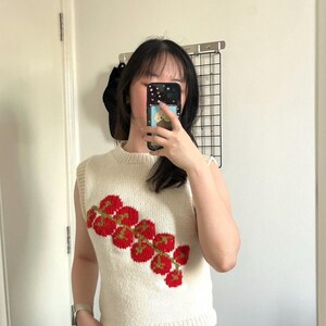 May include: Cream-colored, sleeveless knit top with a red and green floral design. The top has a ribbed hem and neckline. The floral pattern is composed of red circular shapes with green stems. The top is made of a soft, textured yarn.
