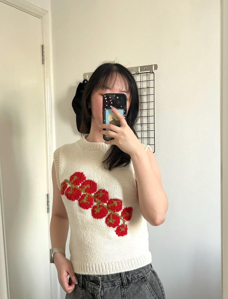 May include: Cream-colored, sleeveless knit top with a red and green floral design. The top has a ribbed hem and neckline. The floral pattern is composed of red circular shapes with green stems. The top is made of a soft, textured yarn.