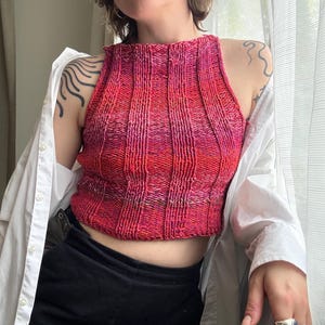 May include: A knitted halter top in shades of red and pink. The top has a ribbed texture and a high neckline. The person is wearing a white button-down shirt and black pants. The top is a handmade garment.