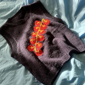 Può includere: Un gilet lavorato a maglia viola scuro con un motivo floreale rosso e giallo. Il top senza maniche è realizzato in un tessuto a maglia strutturato e viene presentato su una superficie azzurra.