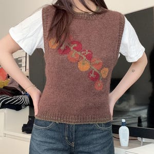 May include: A brown knitted vest with a floral design in shades of red, orange, and yellow. The vest is worn over a white t-shirt and paired with blue denim jeans. The vest has a round neckline and a ribbed hem.