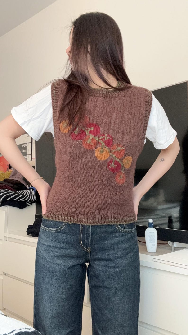 May include: A brown knitted vest with a floral design in shades of red, orange, and yellow. The vest is worn over a white t-shirt and paired with blue denim jeans. The vest has a round neckline and a ribbed hem.