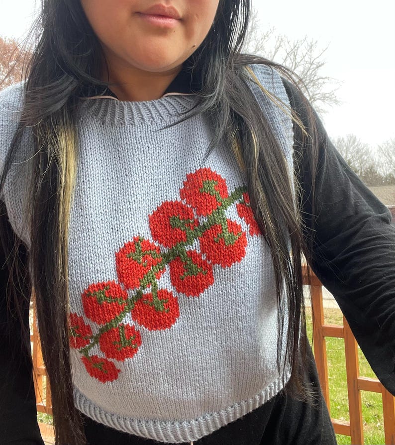 May include: A light blue knitted vest featuring a vibrant tomato design. The tomatoes are red with green stems, creating a colorful contrast against the vest's background. The vest is sleeveless and has a rounded neckline.