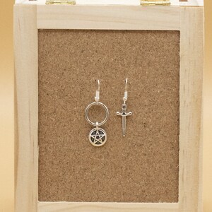 May include: Two silver earrings on a wooden display stand. The earring on the left has a pentagram charm and a circle. The earring on the right has a sword charm.