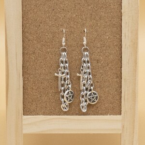 May include: A pair of silver dangle earrings with chain links and charms, including a pentagram and a cross, displayed on a wooden stand.