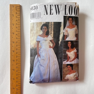 May include: A New Look sewing pattern envelope 6030 for a white wedding dress with an off-the-shoulder neckline and full skirt, shown next to a wooden ruler measuring approximately 24 cm in length, featuring multiple photos of the dress.