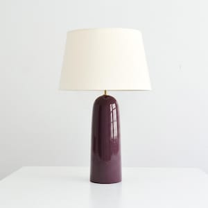 May include: A table lamp featuring a glossy, deep plum-colored ceramic base with a smooth, rounded cylindrical shape. The lamp is topped with a wide, off-white fabric drum shade and includes a small brass-toned metal fitting at the neck.