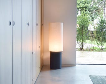 May include: A white cylindrical lampshade with a black base sits on a light gray floor in front of a white wall and a wooden door. The lampshade is illuminated from within, casting a warm glow.