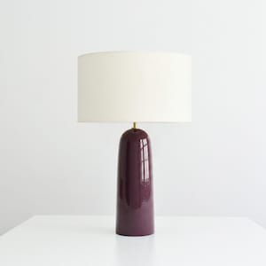 May include: A table lamp featuring a glossy, deep plum-colored ceramic base with a smooth, rounded cylindrical shape. The lamp is topped with a large, off-white fabric drum shade and includes a small gold-toned metal accent at the neck.
