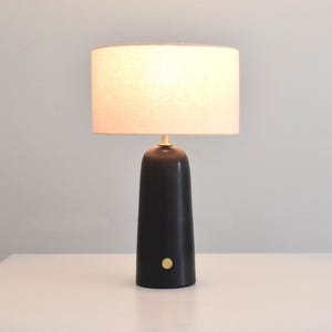 May include: A black ceramic table lamp with a white linen lampshade. The lamp has a gold accent on the top and a gold button on the base.