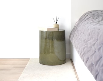 Puede incluir: Una mesa auxiliar cilíndrica de cerámica verde oliva con acabado brillante. Una pila de libros y un pequeño difusor beige con cañas negras encima. La mesa está junto a una cama con una colcha gris acolchada.