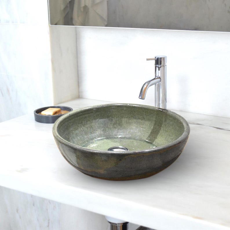 Puede incluir: Un lavabo de cer&aacute;mica redondo y artesanal con un esmalte verde y marr&oacute;n. El lavabo est&aacute; colocado sobre una encimera blanca con un grifo cromado y un peque&ntilde;o jabonera. El di&aacute;metro del lavabo es de unos 40 cm.
