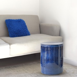 Könnte beinhalten: Eine moderne Wohnzimmerszene mit einem hellgrauen Sofa und einem flauschigen blauen Kissen. Ein zylindrischer blauer Keramik-Beistelltisch mit weißer Oberseite steht neben dem Sofa und setzt einen Farbtupfer in den neutralen Raum.