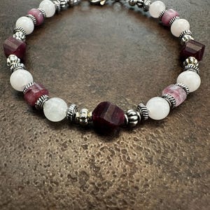 Könnte beinhalten: Ein Armband mit weißen, tiefroten und hellvioletten Edelsteinen. Das Armband hat einen silberfarbenen Verschluss und dekorative Metallperlen. Die Edelsteine sind rund und facettiert. Das Armband ist etwa 18 cm lang.