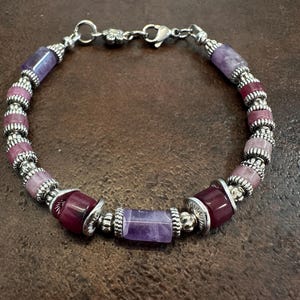 May include: A beaded bracelet featuring a mix of purple and pink gemstones, interspersed with silver-colored metal beads. The bracelet has a lobster clasp closure and a floral accent near the clasp. The gemstones vary in shape and shade.