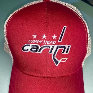 May include: Red trucker hat with a curved brim and mesh back. The hat features the text "Lumpy Head Carini" in white and navy blue, with three white stars above the text. The hat is a classic baseball cap style.