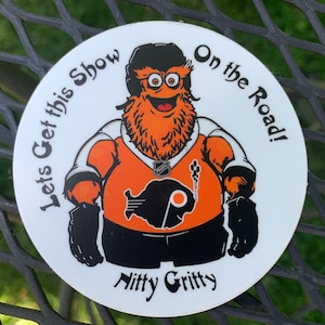 May include: A white round sticker with an orange and black cartoon mascot wearing a hockey jersey. The mascot has a large orange beard and is holding a hockey stick. The text on the sticker reads "Let's Get This Show On the Road!" and "Nitty Gritty".