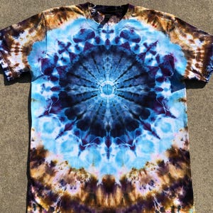 May include: A short-sleeved, V-neck t-shirt with a tie-dye pattern. The shirt features a central design of concentric circles in shades of blue, purple, brown, and white. The tie-dye pattern extends across the entire shirt, creating a unique and colorful design.