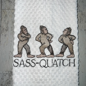 May include: White hand towel with an embroidered design of three Bigfoot figures in various poses. The text "SASS-QUATCH" is embroidered below the figures in black thread. The towel has a textured pattern.