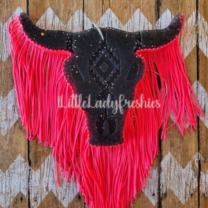 May include: Black and white bull skull with pink fringe. The skull has a geometric design and is decorated with glitter. The fringe is made of yarn and hangs down from the bottom of the skull.