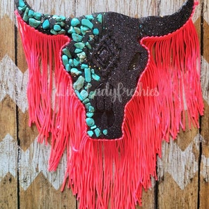 May include: A decorative bull skull with a black glitter finish, adorned with turquoise-colored stones. The skull features bright pink fringe and is mounted on a wooden background with a chevron pattern. The text "Little Lady Freshies" is visible.