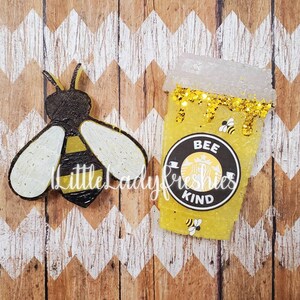 K&ouml;nnte beinhalten: Eine gelbe und schwarze Biene mit wei&szlig;en Fl&uuml;geln und ein gelber und schwarzer Kaffebecher mit goldenen Glitzer und dem Text "Bee Kind" auf einem schwarzen Kreis.