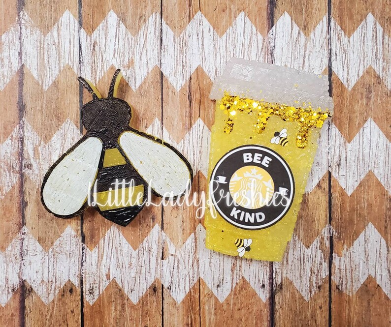 K&ouml;nnte beinhalten: Eine gelbe und schwarze Biene mit wei&szlig;en Fl&uuml;geln und ein gelber und schwarzer Kaffebecher mit goldenen Glitzer und dem Text "Bee Kind" auf einem schwarzen Kreis.