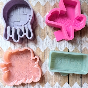May include: Four silicone molds in various shapes and colors. Includes a lavender ghost face, a pink cloud with text, a peach-colored floral design, and a green rectangular mold with text.