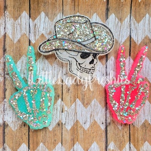 May include: Three glitter peace sign shaped ornaments with a skeleton hand design. The peace signs are in turquoise, pink, and white. The white peace sign has a cowboy hat on top.