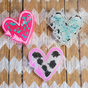 May include: Three resin heart-shaped ornaments with different designs. The first heart is pink with white accents and turquoise glitter. The second heart is white with black accents and blue glitter. The third heart is white with black accents and a cow print design.