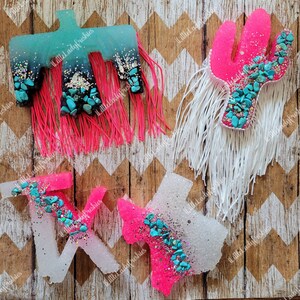May include: Four resin air freshener shapes with turquoise glitter and turquoise stones. The shapes are a cactus, a bird, and two Texas shapes. The cactus and one Texas shape are pink, the bird and the other Texas shape are white.