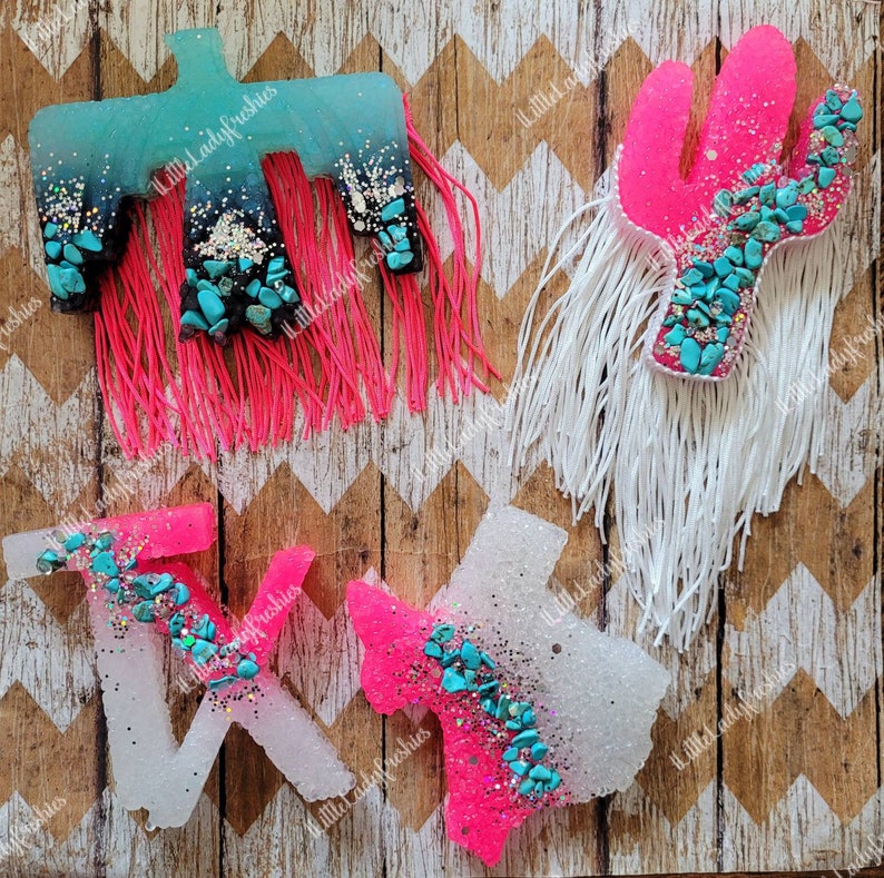 May include: Four resin air freshener shapes with turquoise glitter and turquoise stones. The shapes are a cactus, a bird, and two Texas shapes. The cactus and one Texas shape are pink, the bird and the other Texas shape are white.