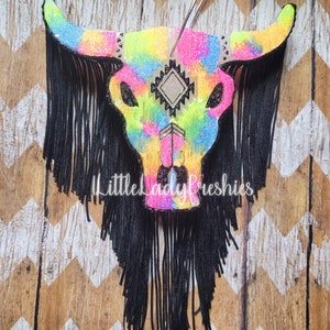 May include: A colorful tie-dye skull with black fringe hanging from the bottom. The skull has a geometric design in the center and is made of glitter.
