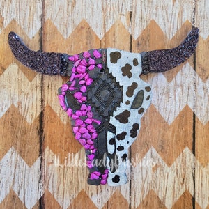 May include: A black and white cow skull with pink and black mosaic details. The skull is made of a textured material and has a glittery finish.