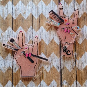 May include: Two pink glitter resin hands making the rock and roll hand gesture. One hand is holding a set of hairdressing tools, the other is holding makeup brushes.