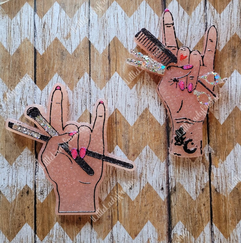 May include: Two pink glitter resin hands making the rock and roll hand gesture. One hand is holding a set of hairdressing tools, the other is holding makeup brushes.