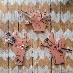 May include: Three pink resin hand-shaped ornaments with black outlines. Each hand is making a rock and roll hand gesture. The hands are holding different beauty tools, including makeup brushes, a comb, and a mascara wand. The ornaments are decorated with glitter.