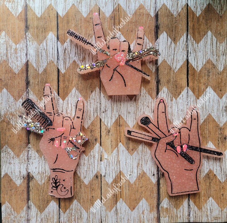 May include: Three pink resin hand-shaped ornaments with black outlines. Each hand is making a rock and roll hand gesture. The hands are holding different beauty tools, including makeup brushes, a comb, and a mascara wand. The ornaments are decorated with glitter.