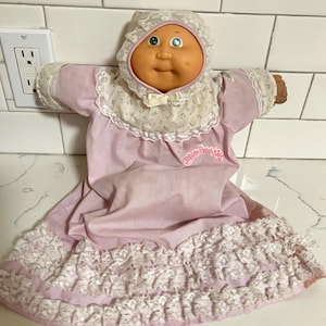 May include: A vintage Cabbage Patch Kids doll wearing a light pink dress with white lace trim and a matching bonnet. The doll has a round face, blue eyes, and a small bow. The doll's dress has the words "Cabbage Patch Kids" on the front.
